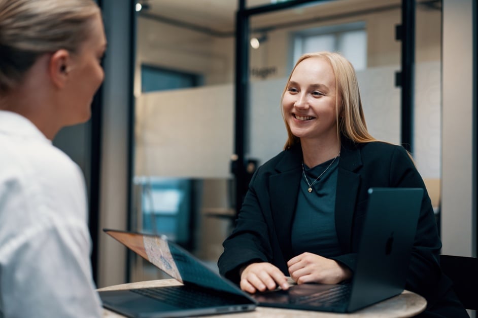 Woman with blonde hair and black blazer smiling and looking to the left while resting hands on laptop